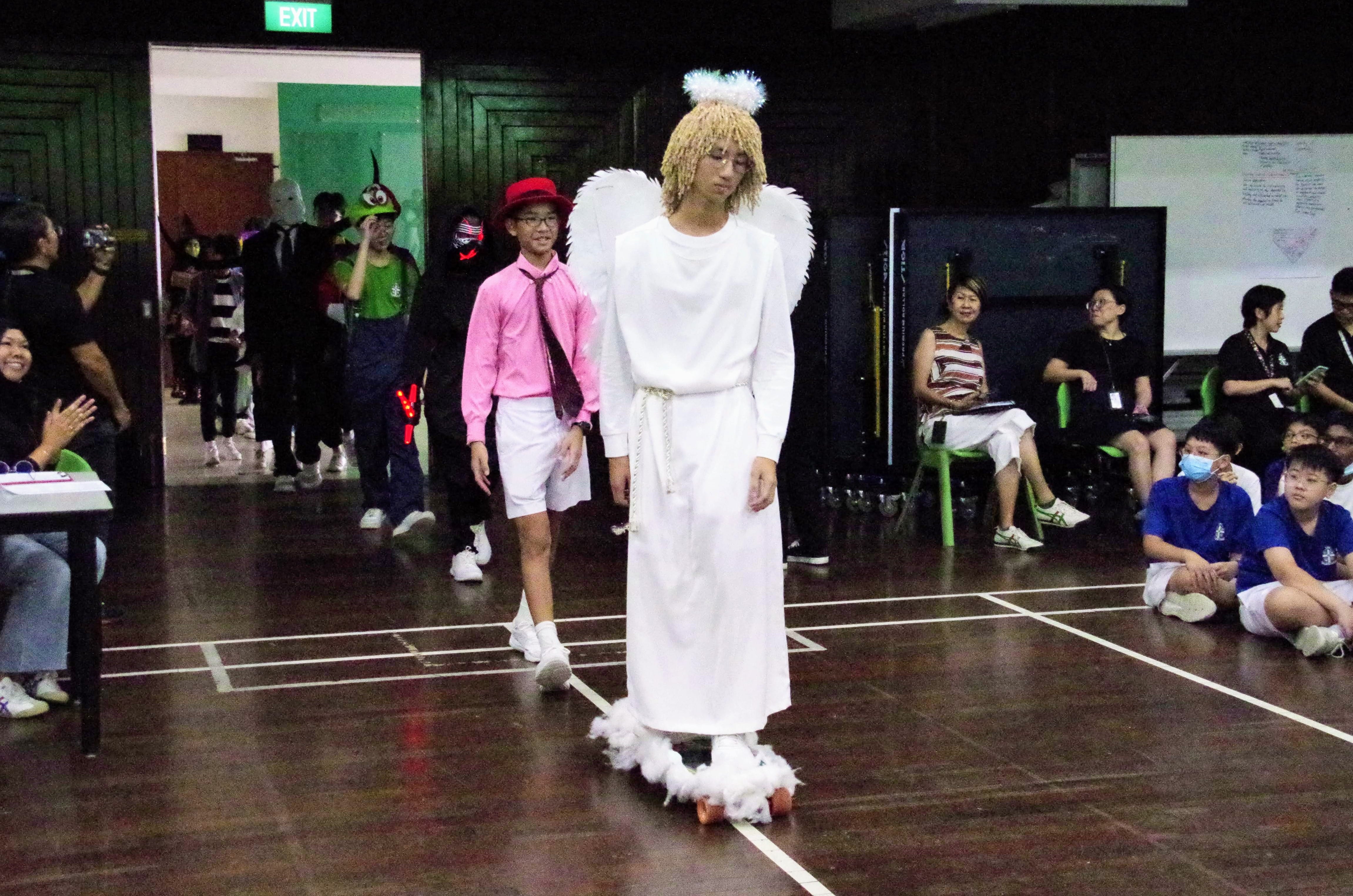 A student dressed as an angel in a white robe with wings and a halo, roller-skating indoors during a costume parade.